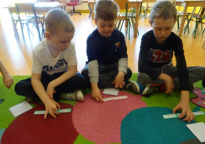 Trzech chłopców siedzi na dywanie i układa zdania z rozsypanek wyrazowych.